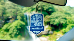 Fresh Falls car air freshener badge hanging from rearview mirror with waterfall scenery and green forest background visible through windshield.