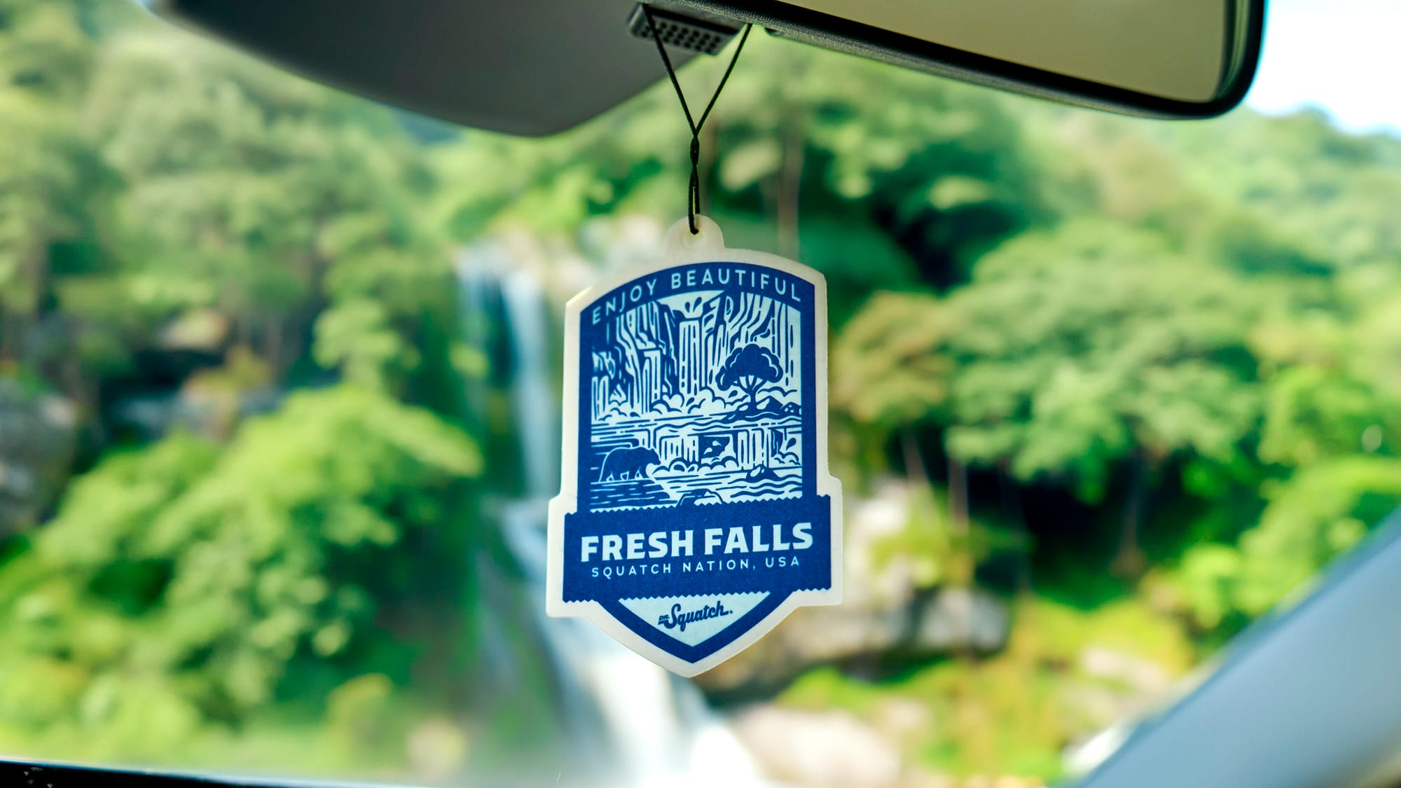 Fresh Falls car air freshener badge hanging from rearview mirror with waterfall scenery and green forest background visible through windshield.