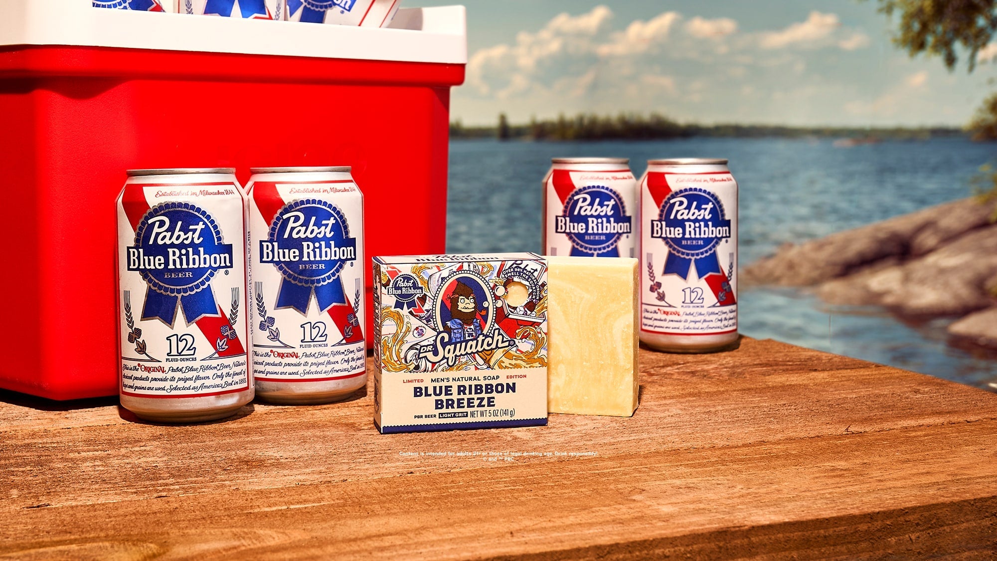 Pabst Blue Ribbon beer products displayed outdoors on a wooden table by the water, including four cans, a red cooler, and a blue ribbon breeze soap box